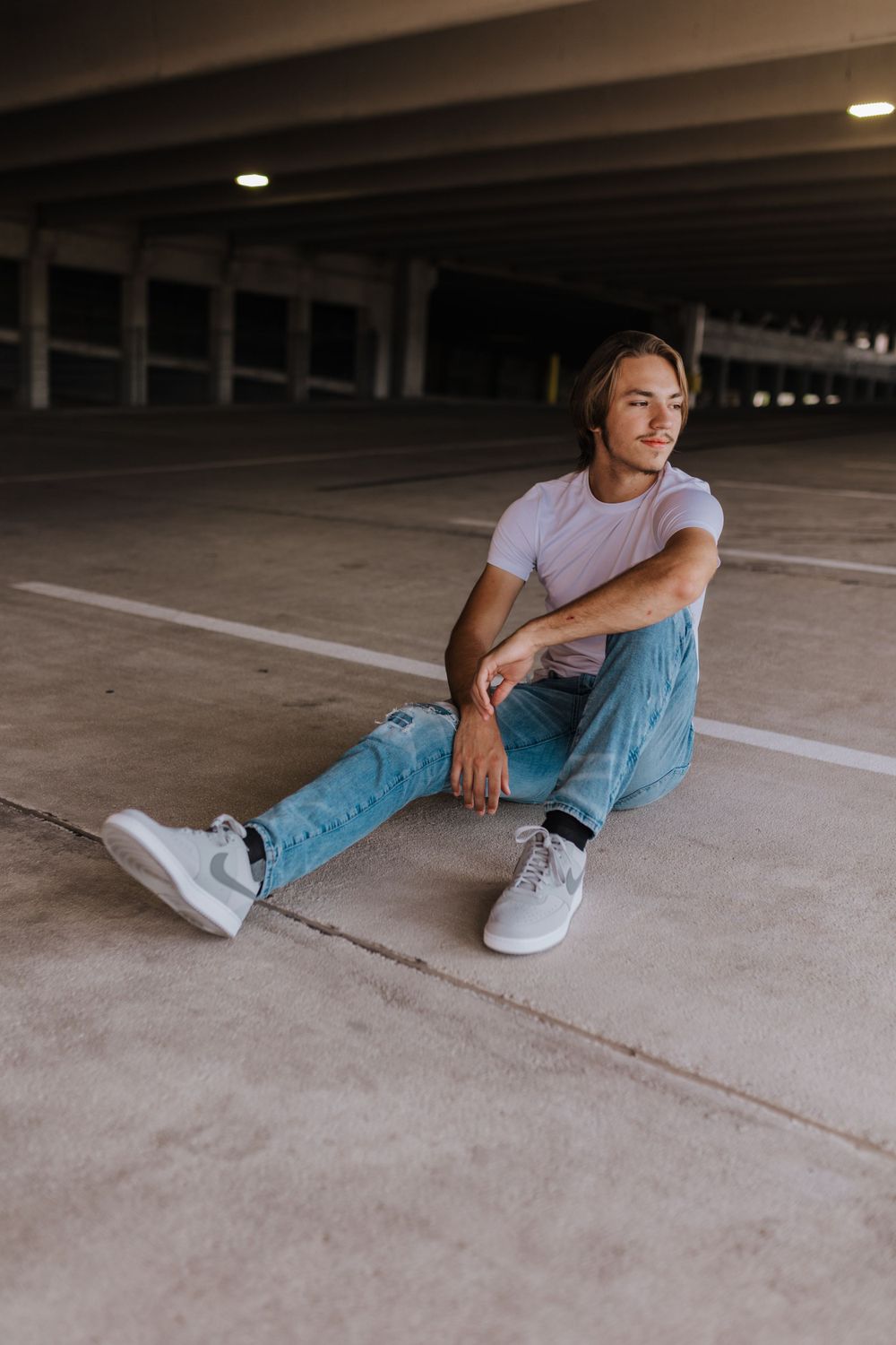 A casual portrait of someone sitting on a parking garage floor wearing light wash jeans and white sneakers.