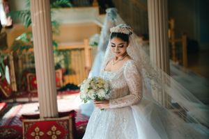 Elegant bride in long-sleeved lace wedding gown holding white bouquet in ornate interior.