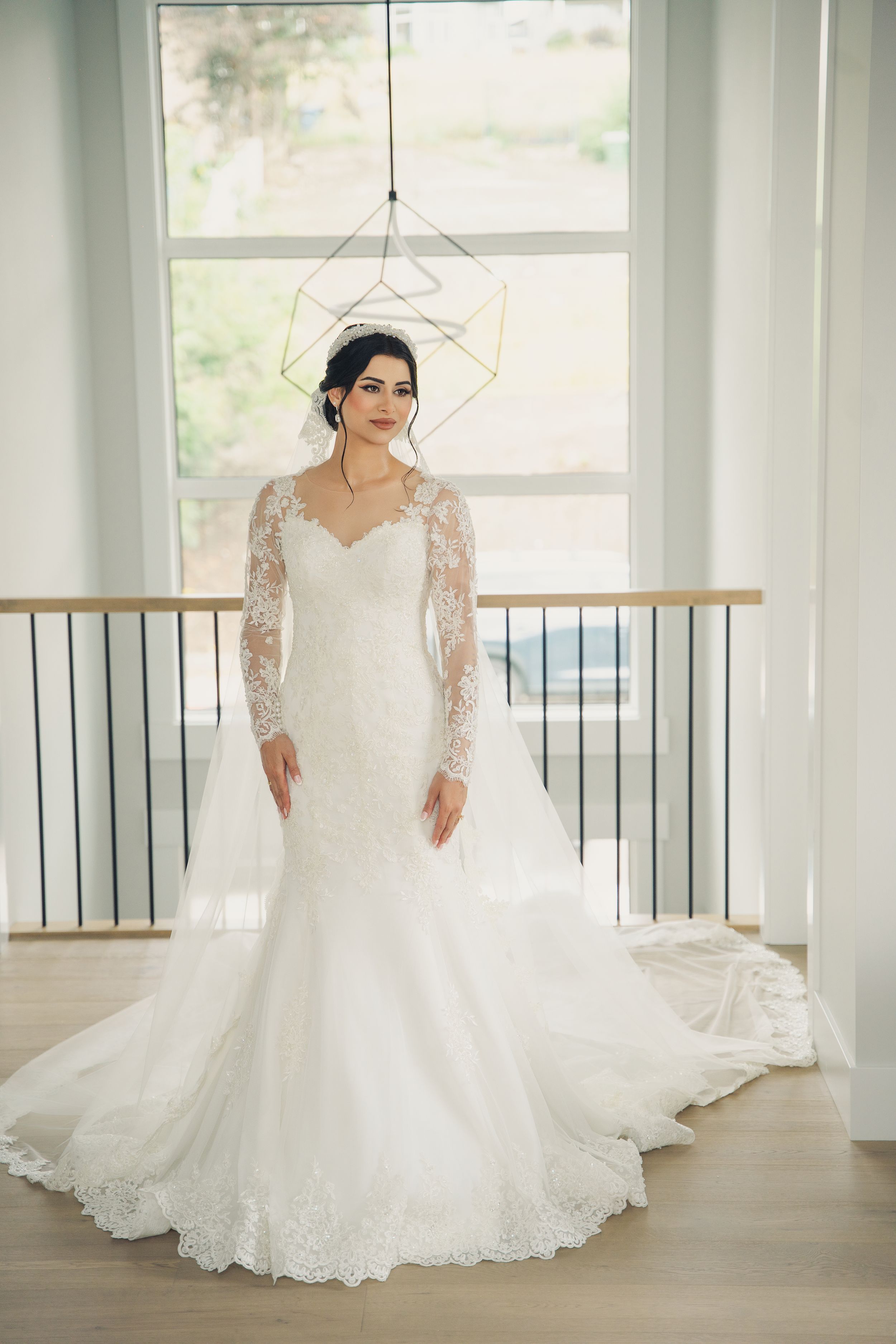 Elegant bride in lace long-sleeve wedding gown poses beneath geometric light fixture.