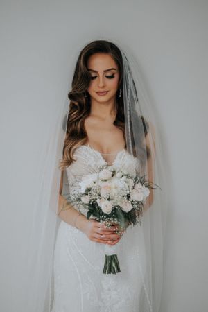 Classic bridal portrait with flowing veil and white rose bouquet against neutral background.