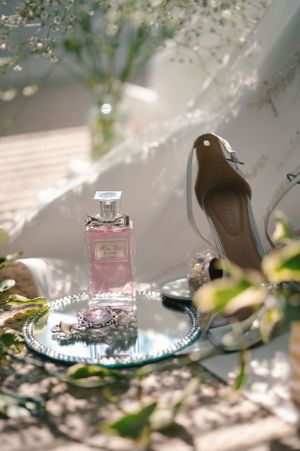 Pink perfume bottle and bridal shoe displayed on reflective surface with delicate floral elements.