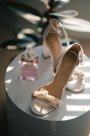 Wedding accessories including elegant white heels and pink perfume bottle arranged in soft natural light.