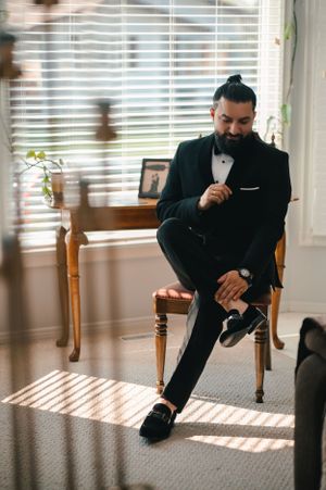 Groom in classic tuxedo sitting on wooden chair by window with natural light streaming in.