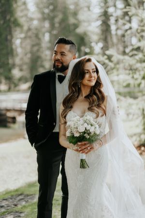 Elegant bride in lace gown holds white bouquet while groom in black tuxedo stands in forest setting.