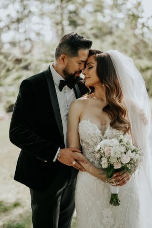 Intimate wedding photo of couple sharing romantic moment in forest with bride holding white bouquet.