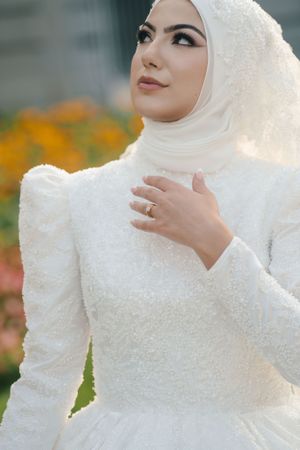 Close up of bride in white hijab wedding dress showing engagement ring amidst orange flowers.