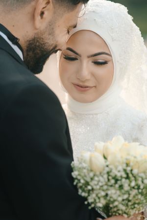 Intimate moment between newlyweds with bride in white hijab and bouquet of cream roses.