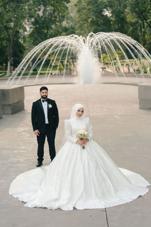 Newlyweds pose in front of decorative water fountain with bride in traditional white hijab and ballgown.