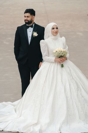 Muslim bride in modest white ballgown and groom in tuxedo pose for formal wedding portrait.