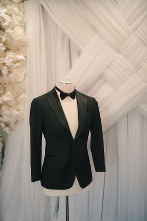 Classic black tuxedo jacket with bow tie displayed on mannequin against white draped backdrop.