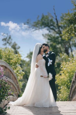 Romantic wedding couple standing on bridge against blue sky and green foliage.