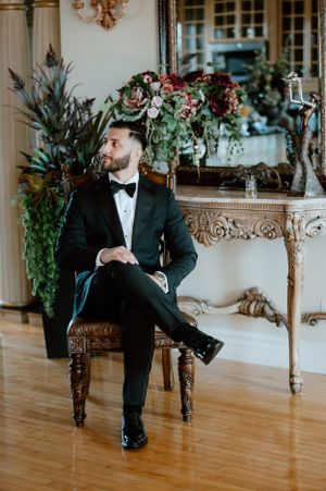 A groom in classic tuxedo sits elegantly on an antique chair near ornate floral arrangements.