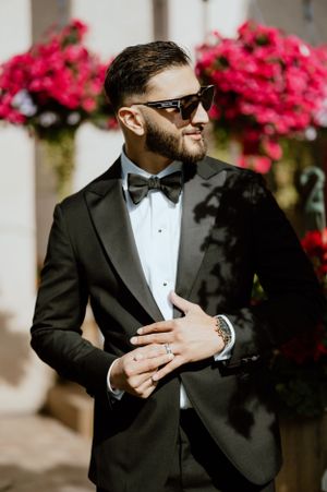 Stylish groom in sunglasses and tuxedo poses near vibrant pink flowers.