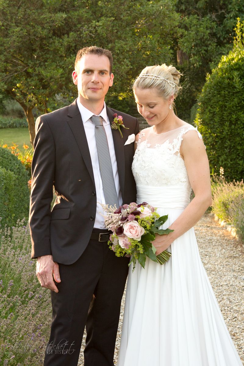Newlywed couple poses in a garden setting with bride holding pink and purple bouquet.