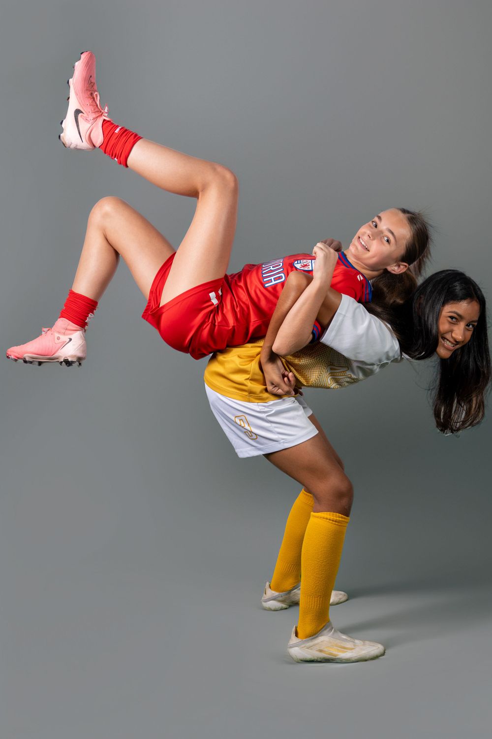Two soccer players in red and yellow uniforms pose playfully with one balanced on the other's back against a gray backdrop.