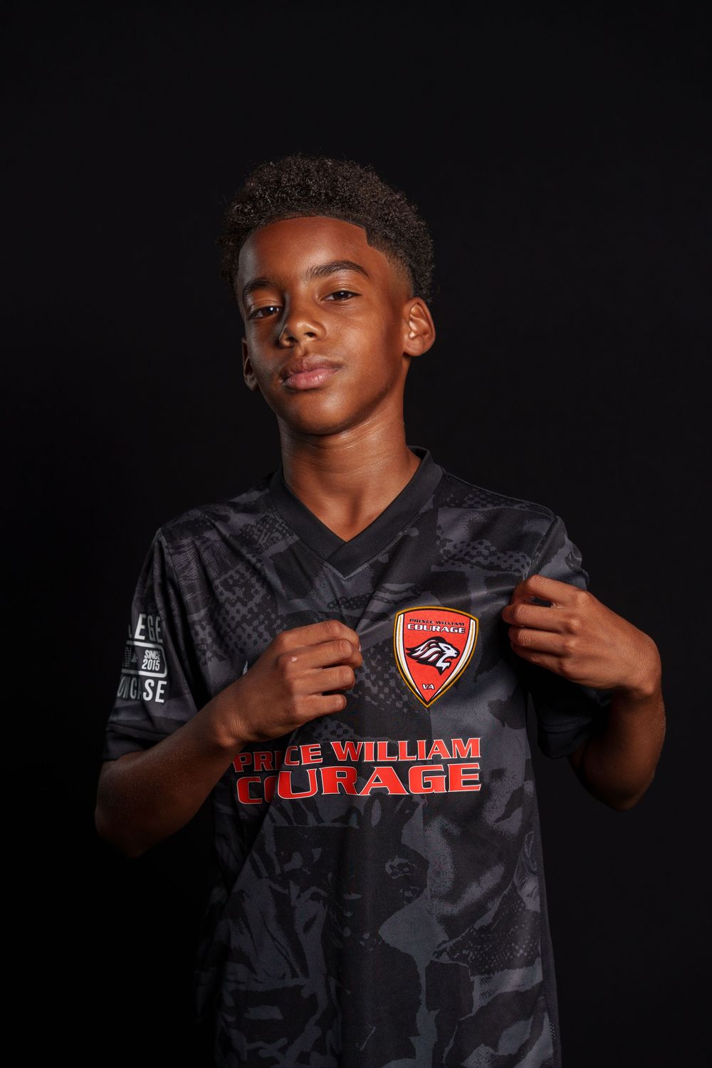 Young athlete poses in dark jersey with The William Courage team logo against black background.