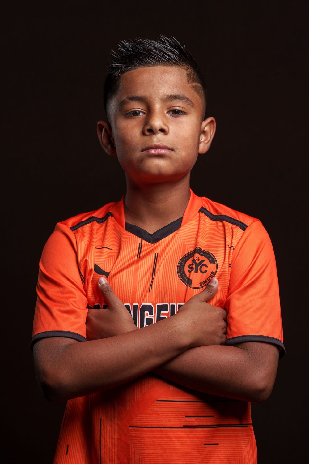 Youth soccer player poses in bright orange jersey against dark background in dramatic portrait series.