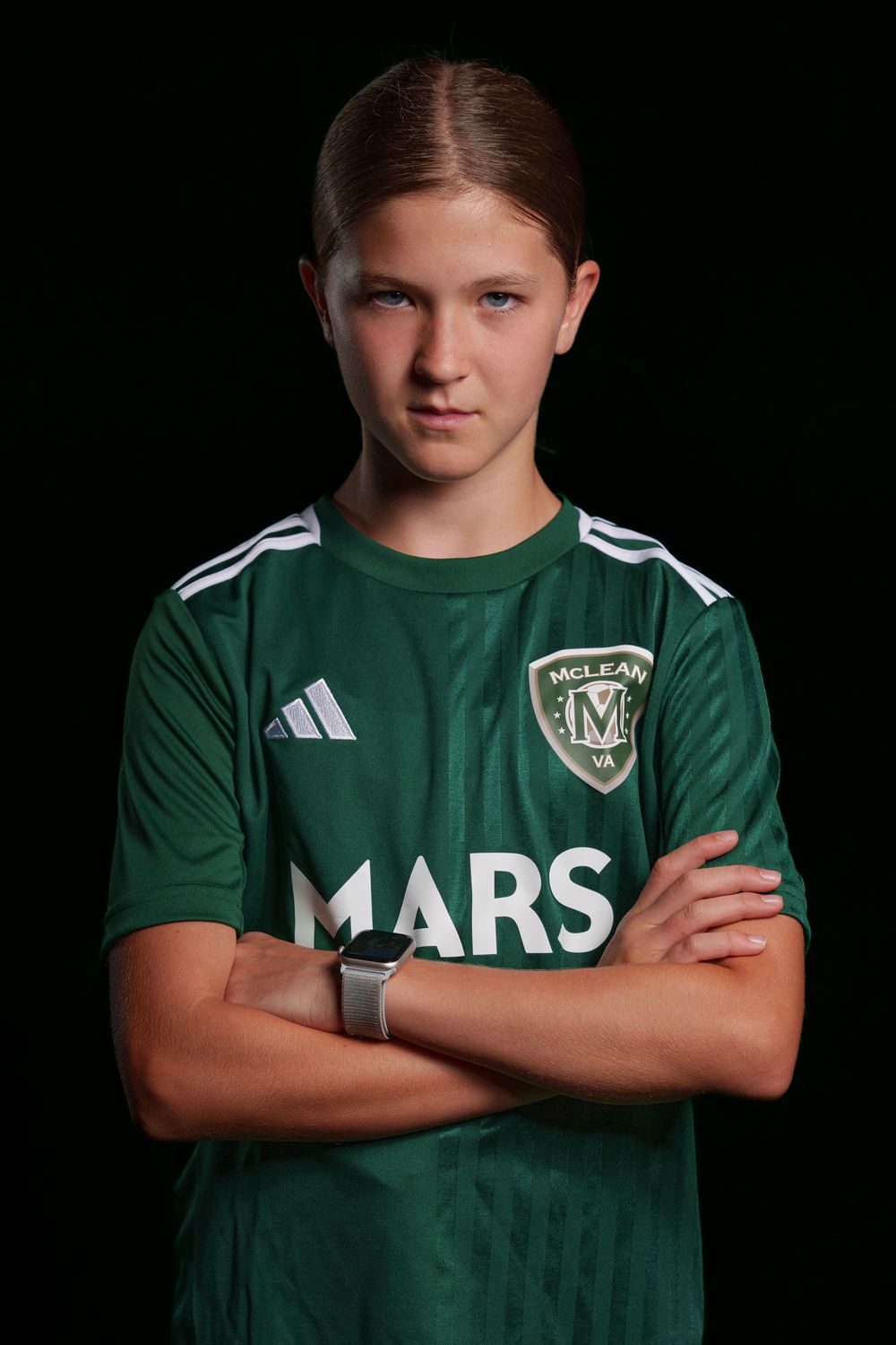 Youth soccer player wearing green Mars team jersey poses with arms crossed against dark background.