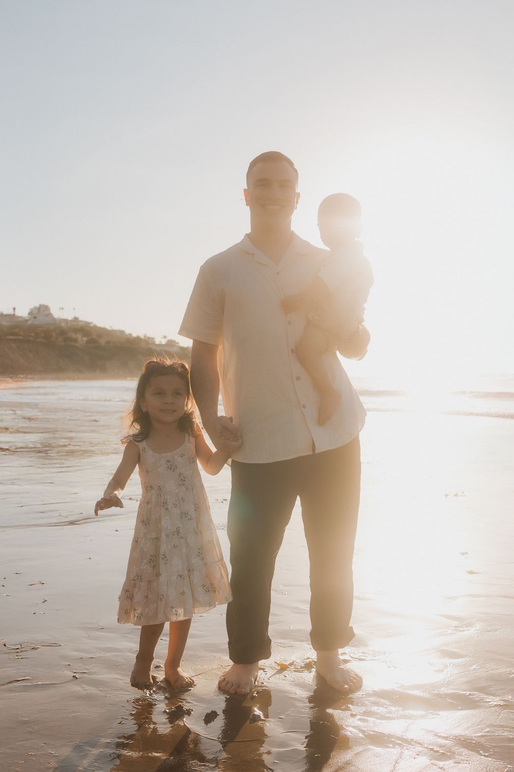 Father holds hands with children while standing in shallow ocean waves at sunset.