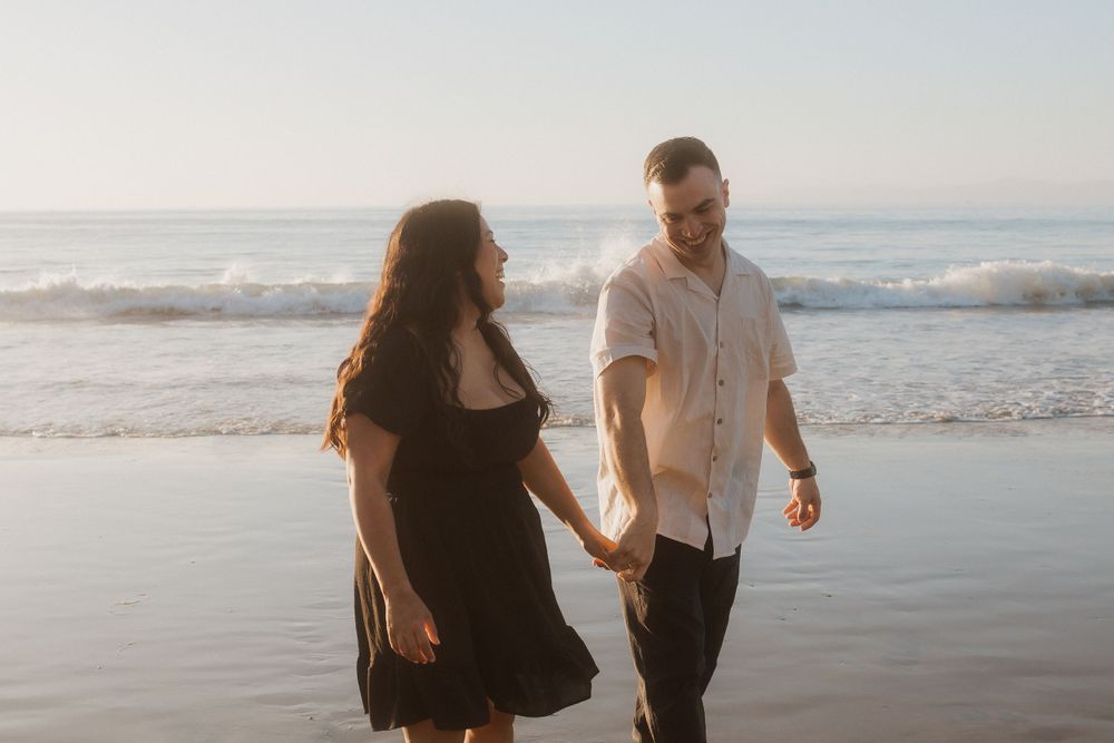 A couple walking together on a sunny beach with waves crashing behind them.