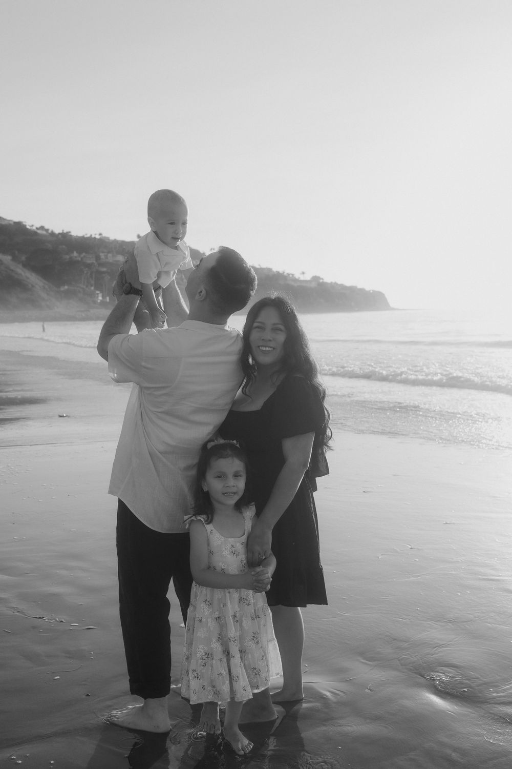 A black and white family portrait on the beach with cliffs in the background.