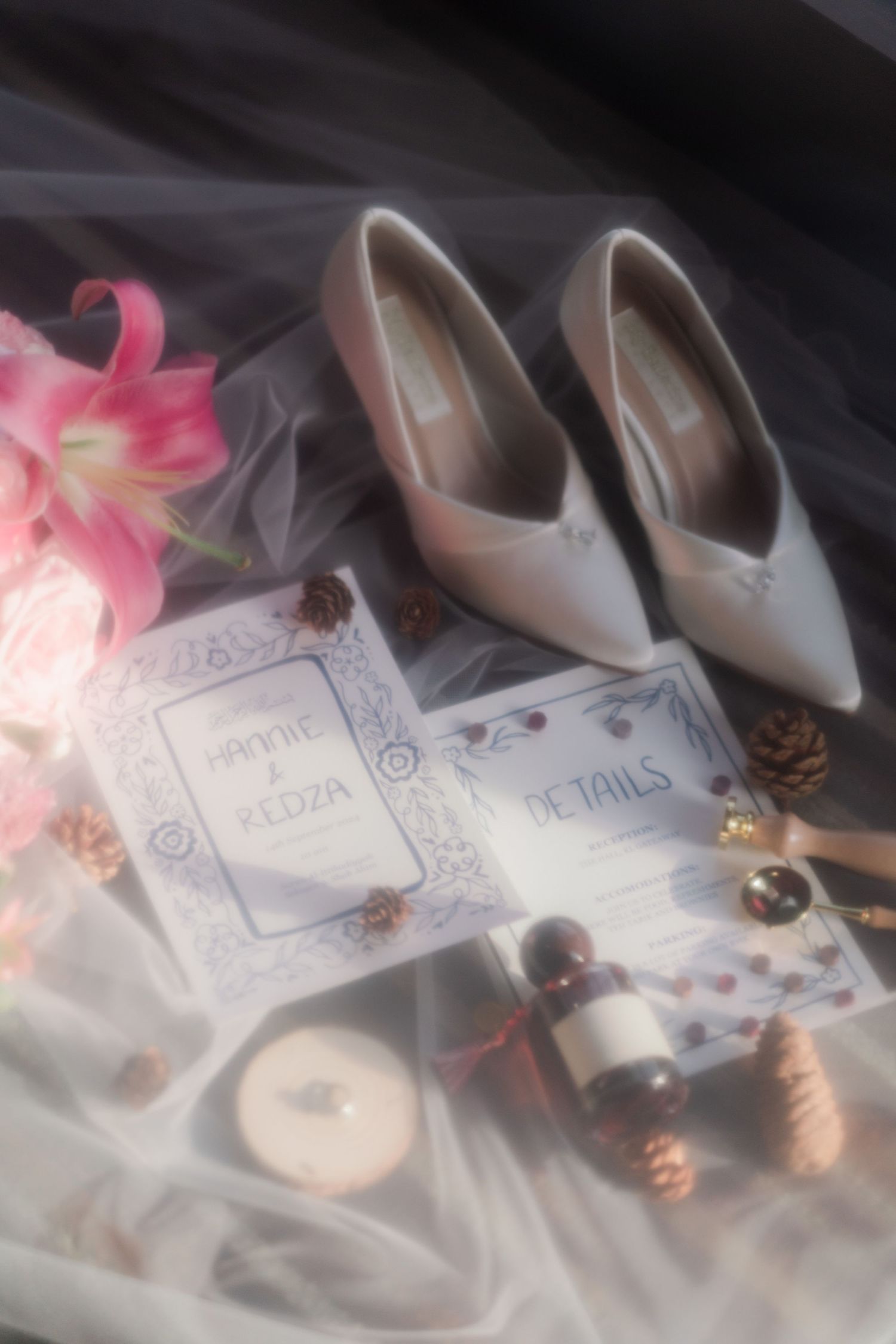 Wedding details flatlay with pink flowers, beige heels, and invitation cards on tulle fabric.