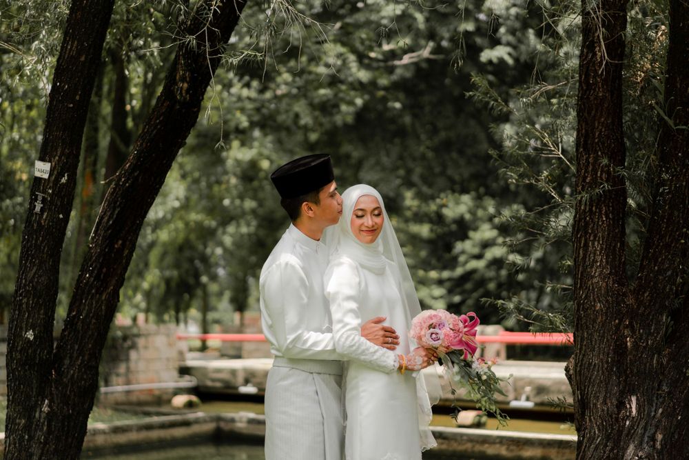 A Muslim couple in traditional white wedding attire share a moment in a park setting.