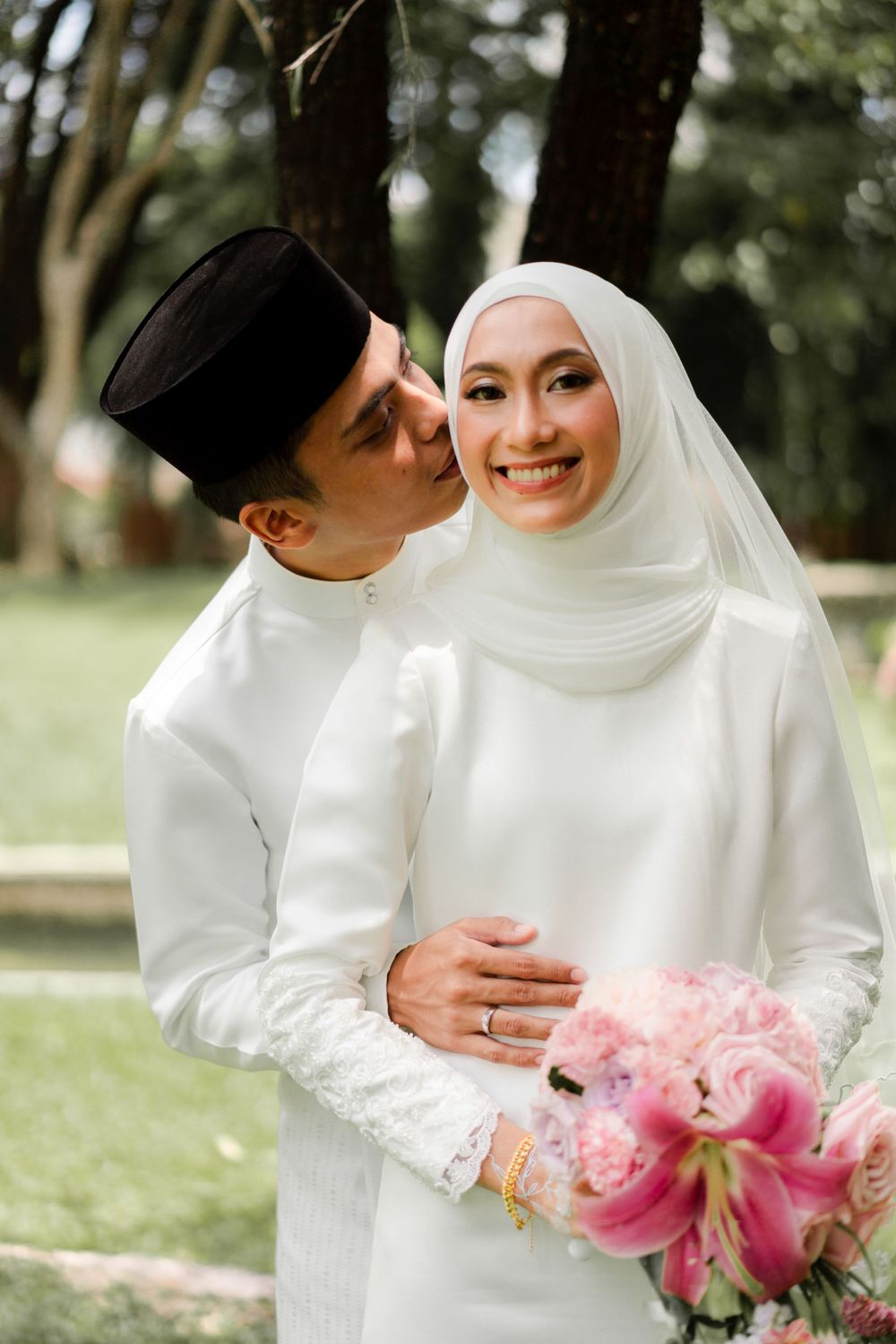 A traditional Muslim wedding couple in white attire share a tender moment with pink flowers.