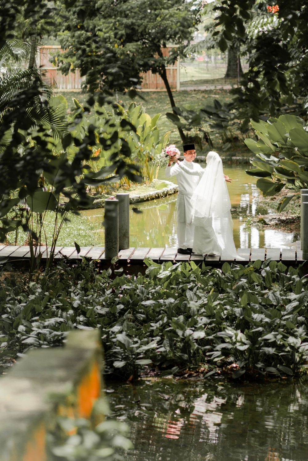 A couple in white stands on a wooden bridge in a lush garden setting during their wedding.