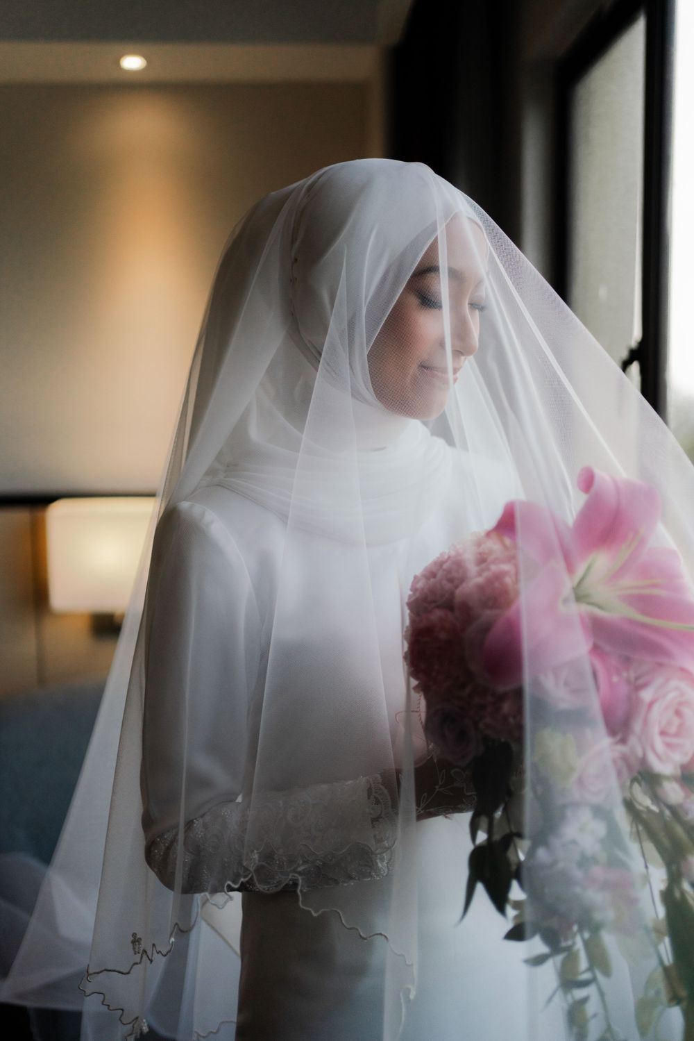 A bride in white hijab and dress holds pink flowers while standing by a window.