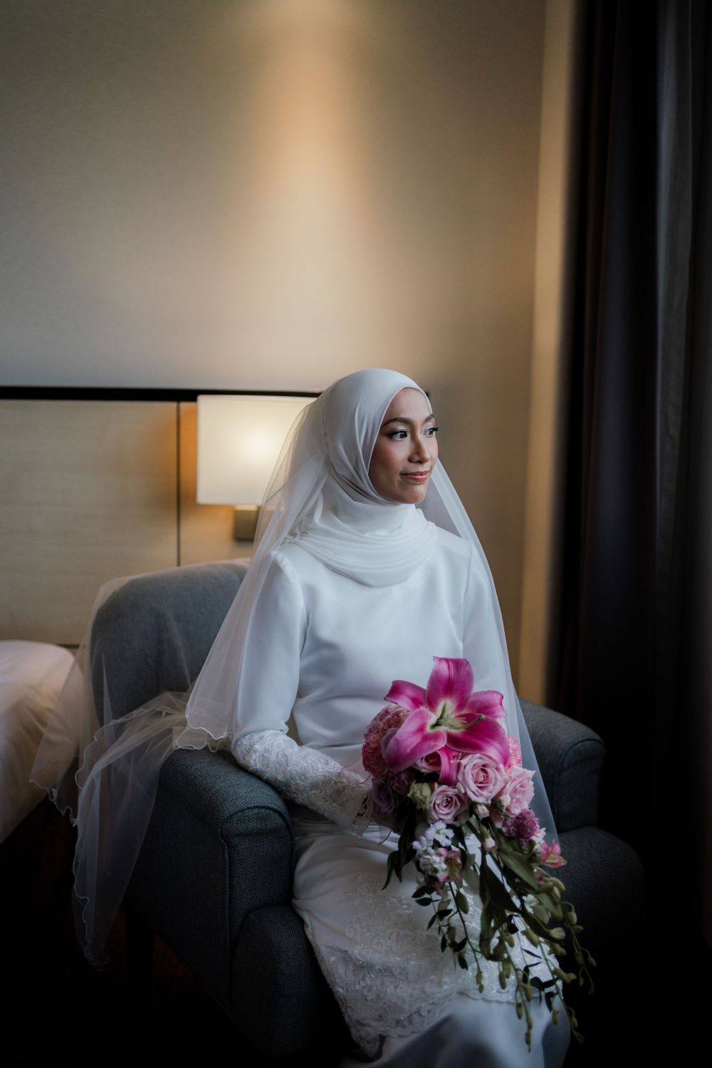 Serene bride in white hijab and dress holding pink floral bouquet while seated in hotel room.