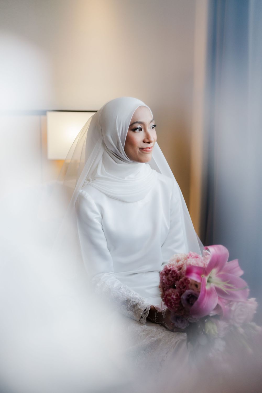 Elegant bride wearing white hijab and dress poses with pink flowers by window.