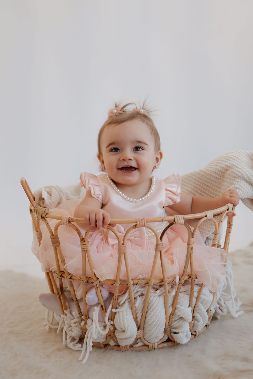 Baby in pink dress sitting in decorative wicker basket against white background.