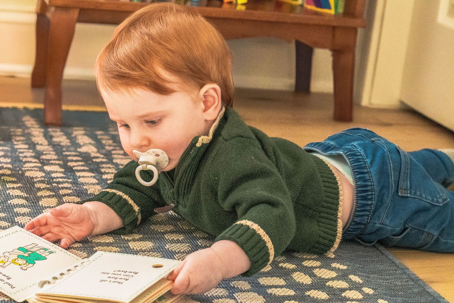 A child in a green sweater lies on a patterned rug reading a book.