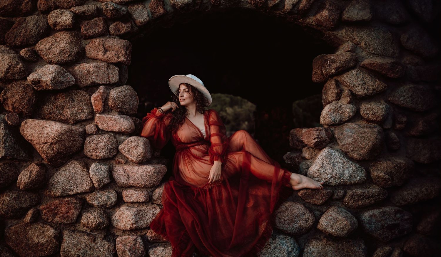 Woman in flowing red dress poses dramatically against ancient stone wall architecture.