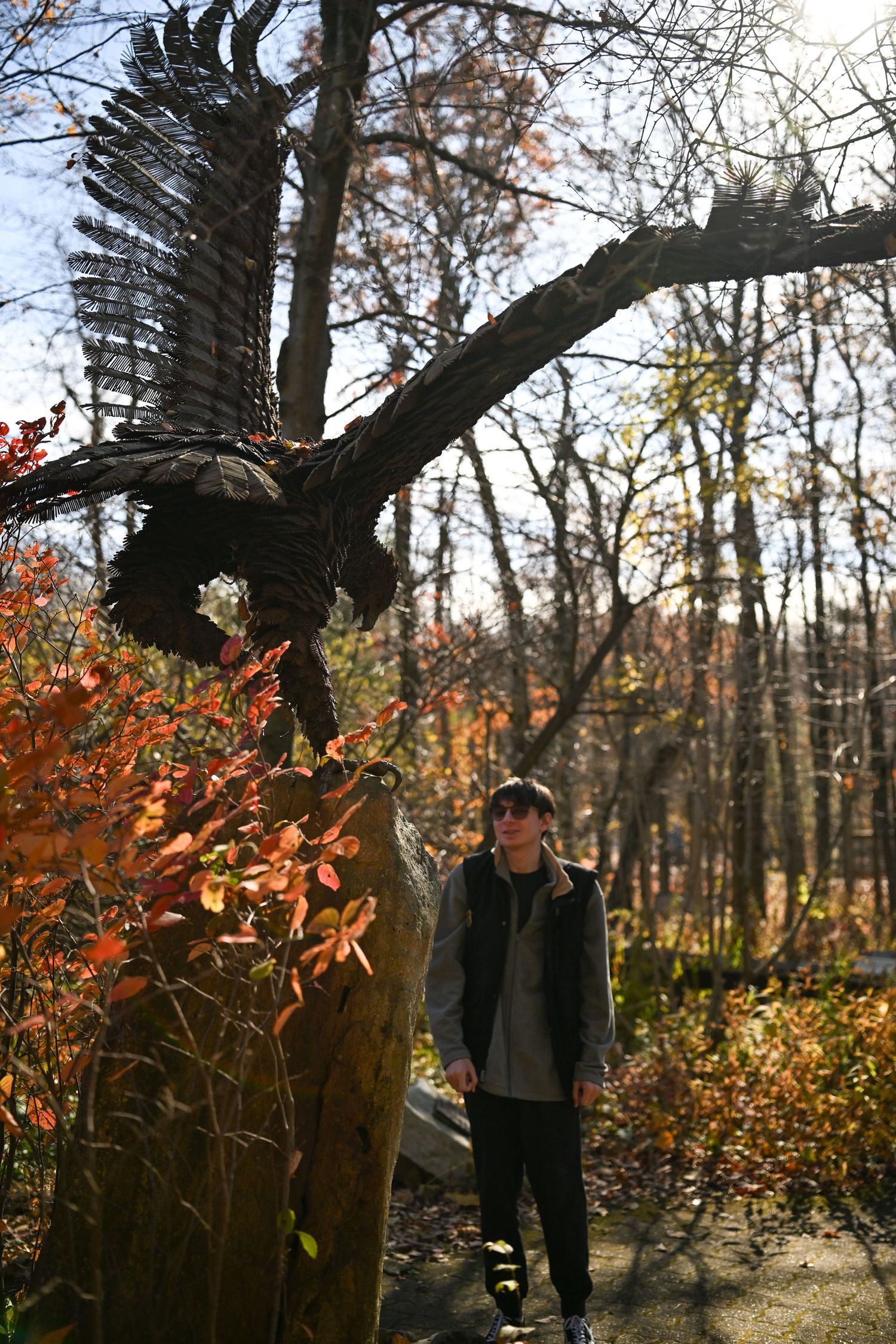 Photograph of a young man candidly posing in front of a large metal sculpture of a bird in flight