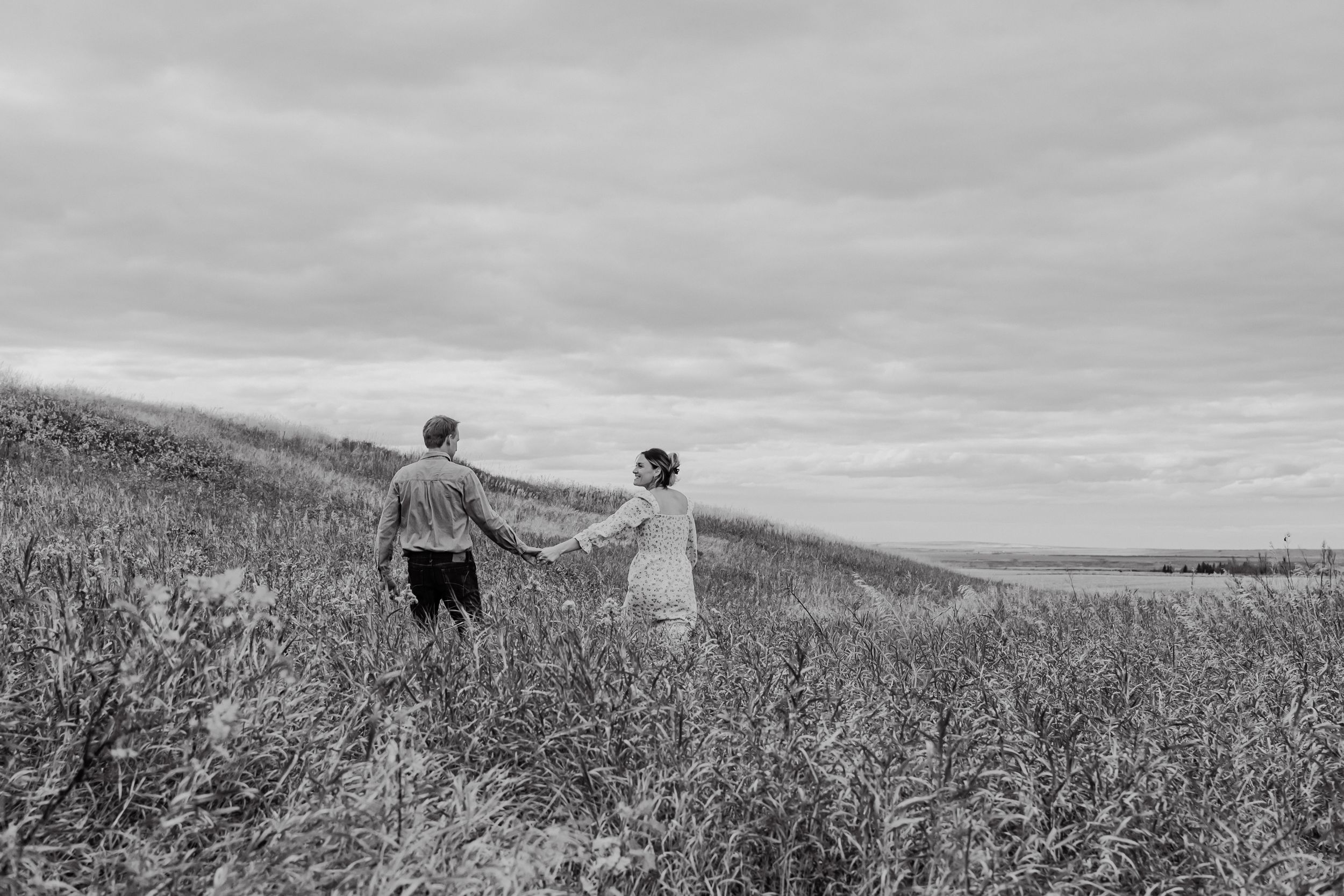 Black and white photos showing a couple walking through a field of tall grass and flowers on a cloudy day.