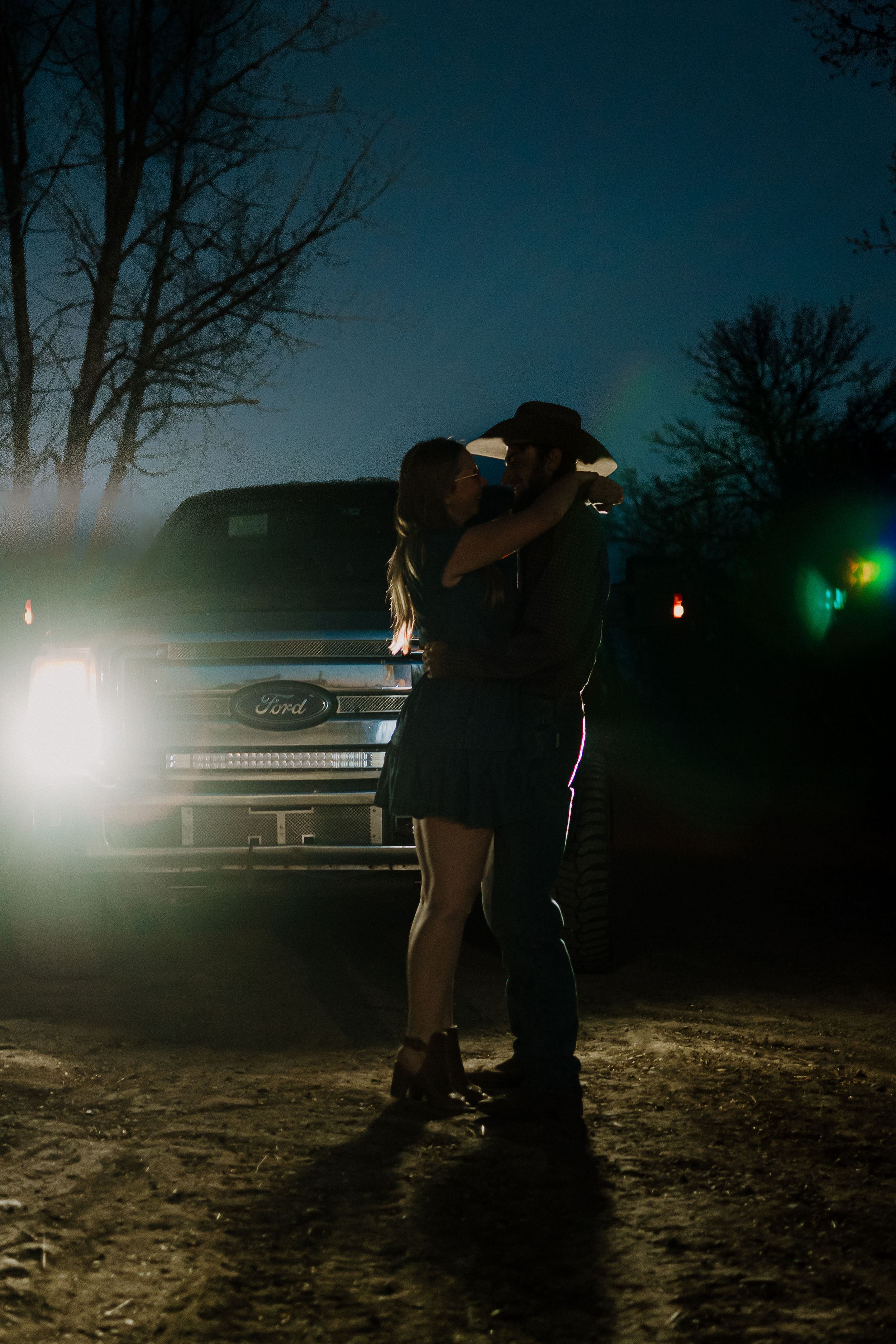 Silhouettes of people standing near a vintage car with headlights on at night.
