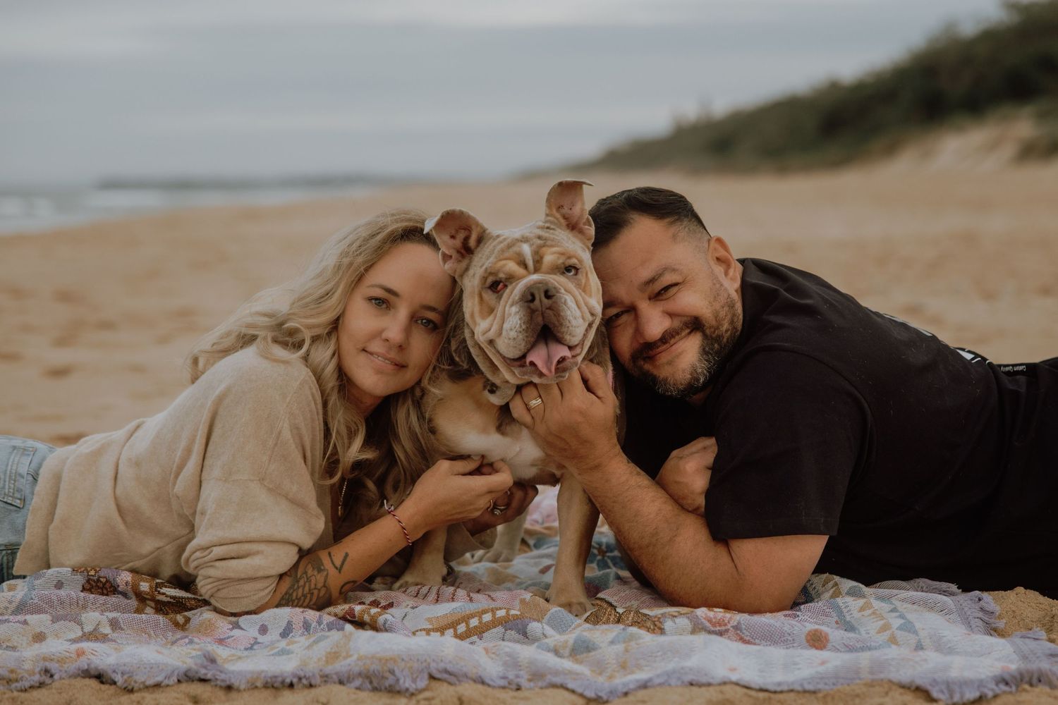 A couple lies on a beach blanket with their English Bulldog during a cloudy coastal photoshoot.