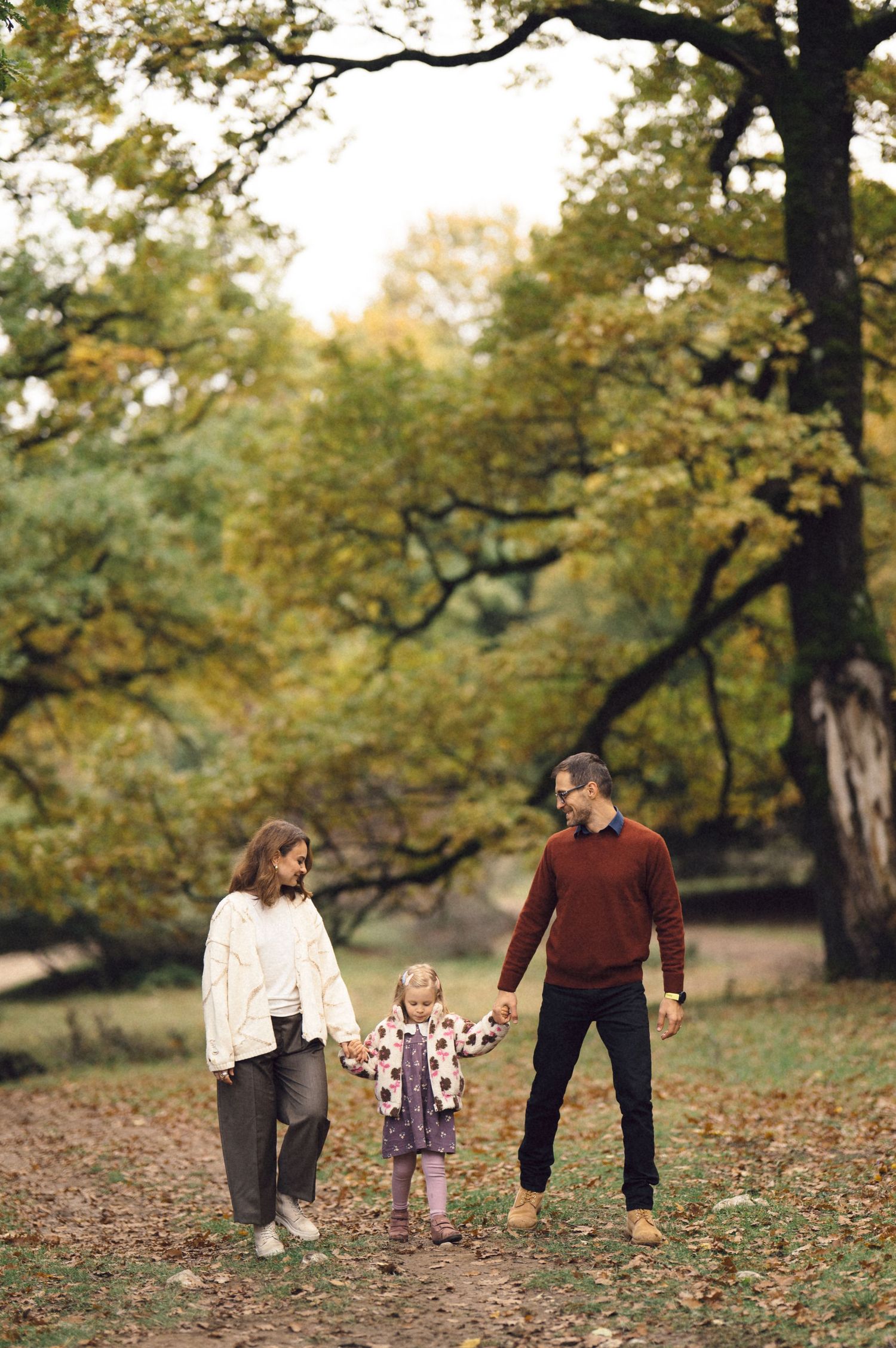 A family walks together through a park on an autumn day with fallen leaves scattered on the ground.