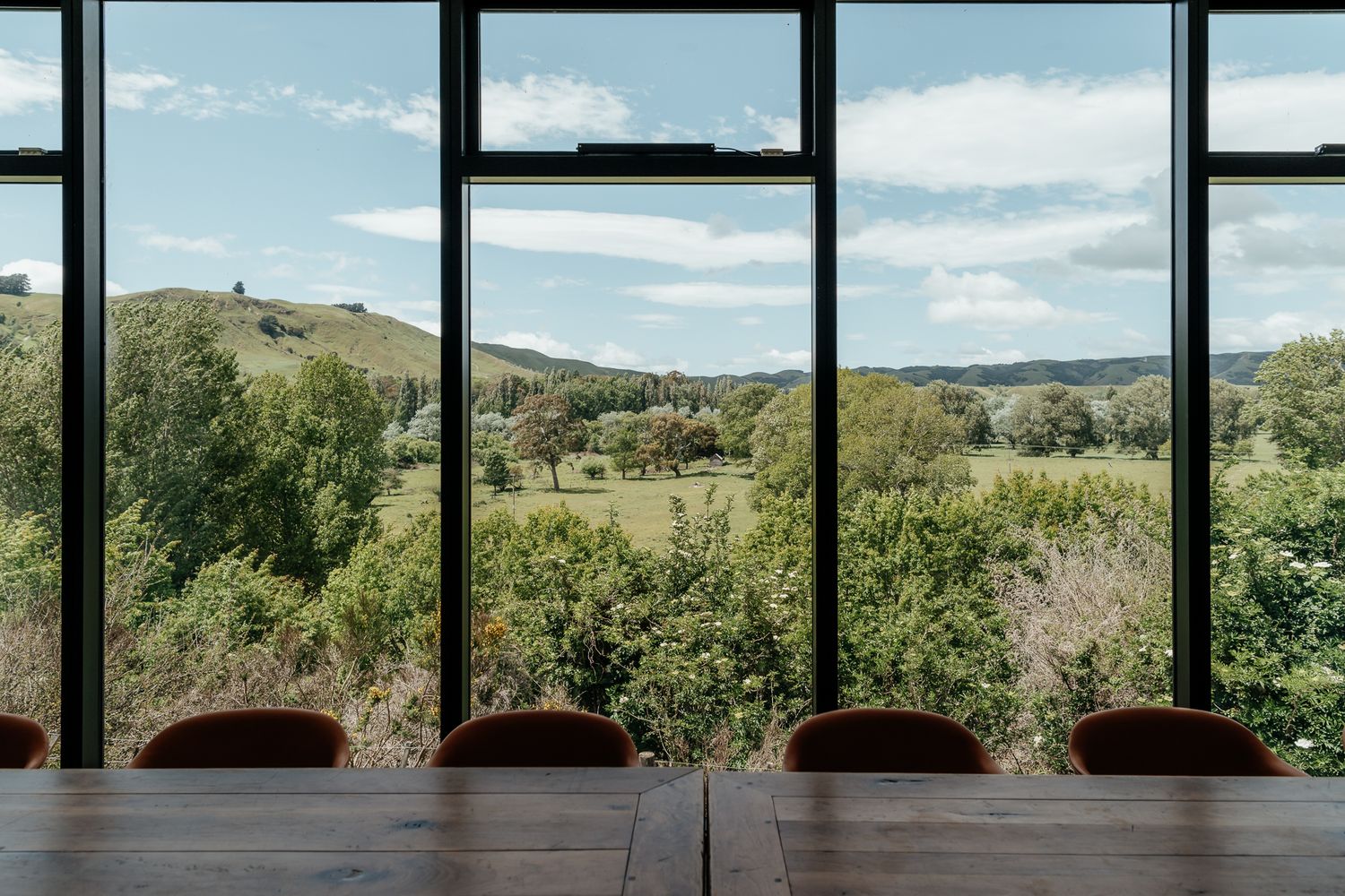 Panoramic view through large windows showing rolling green hills and trees under a bright blue sky with scattered clouds by August and Vine