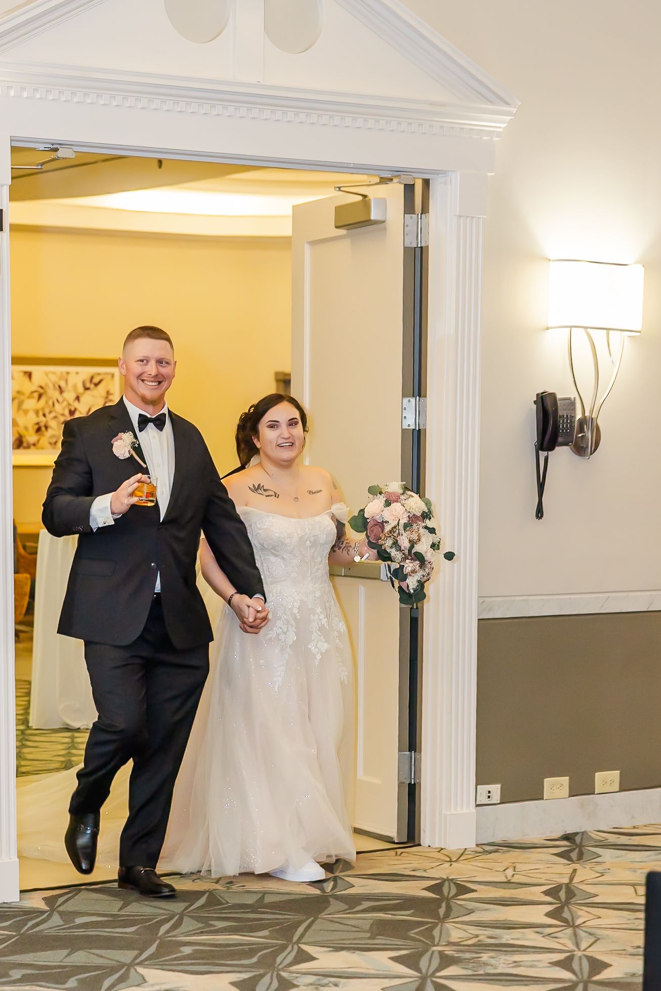 A wedding couple poses playfully behind white columns in a reception venue with decorative carpet.