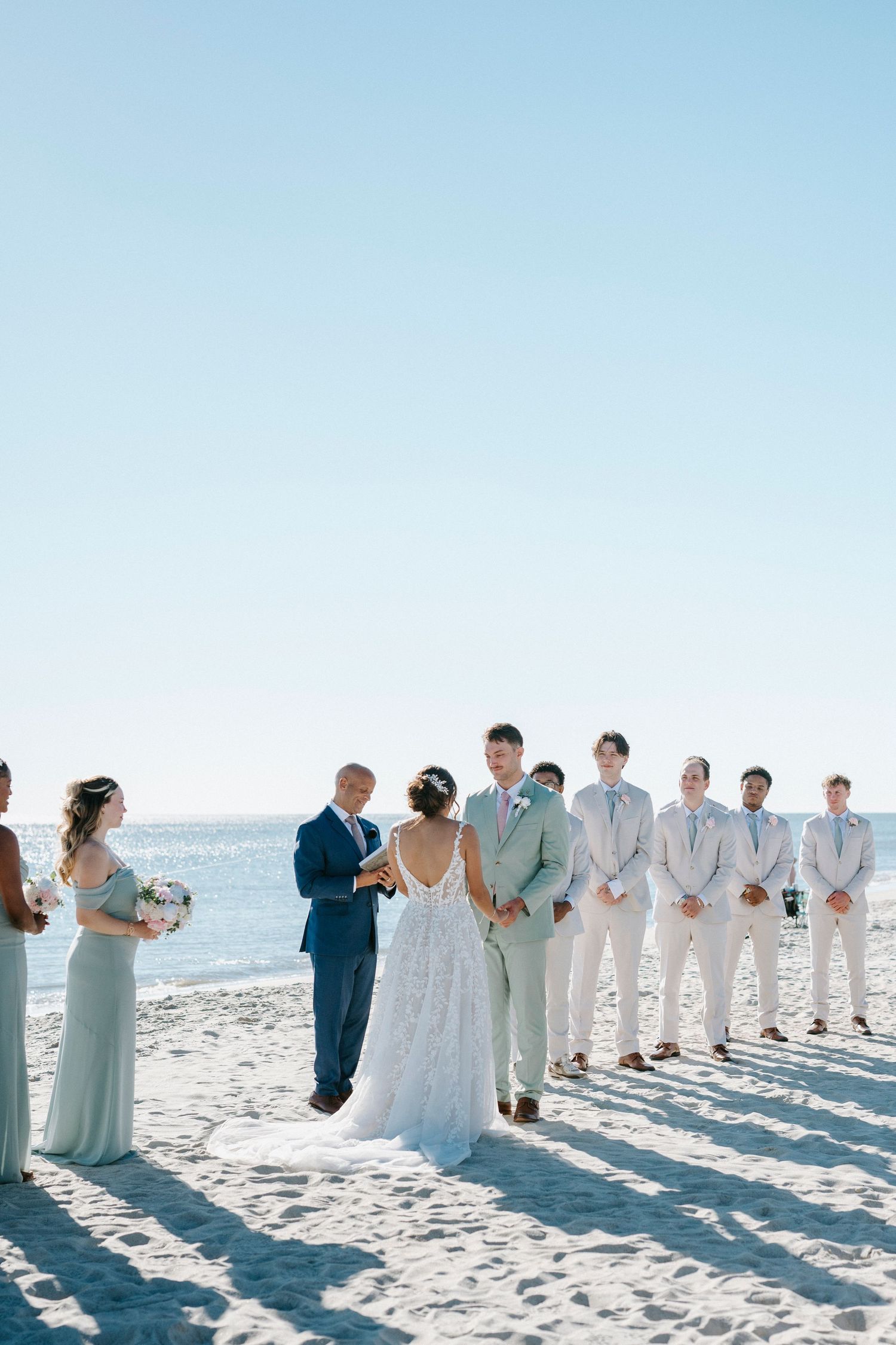 Wedding party in formal attire stands in a line casting long shadows on a sunny beach with blue sky.