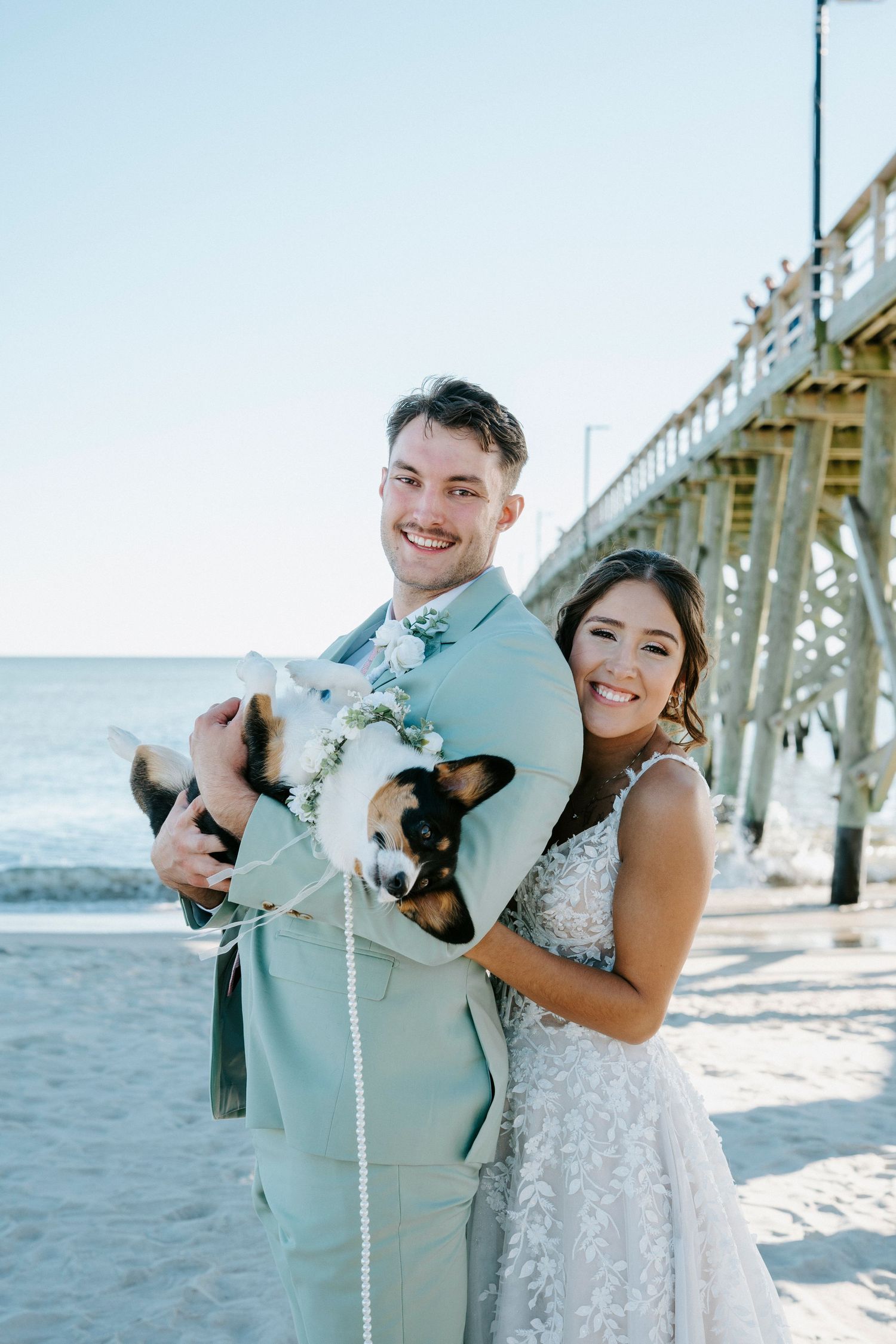 A couple shares joyful moments with their small dog at a beach pier on a sunny day.