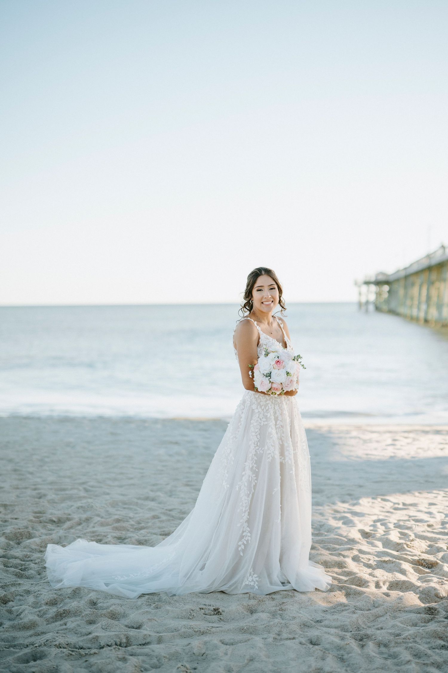 A person in a flowing white wedding dress stands gracefully on a sandy beach at sunset near the ocean.
