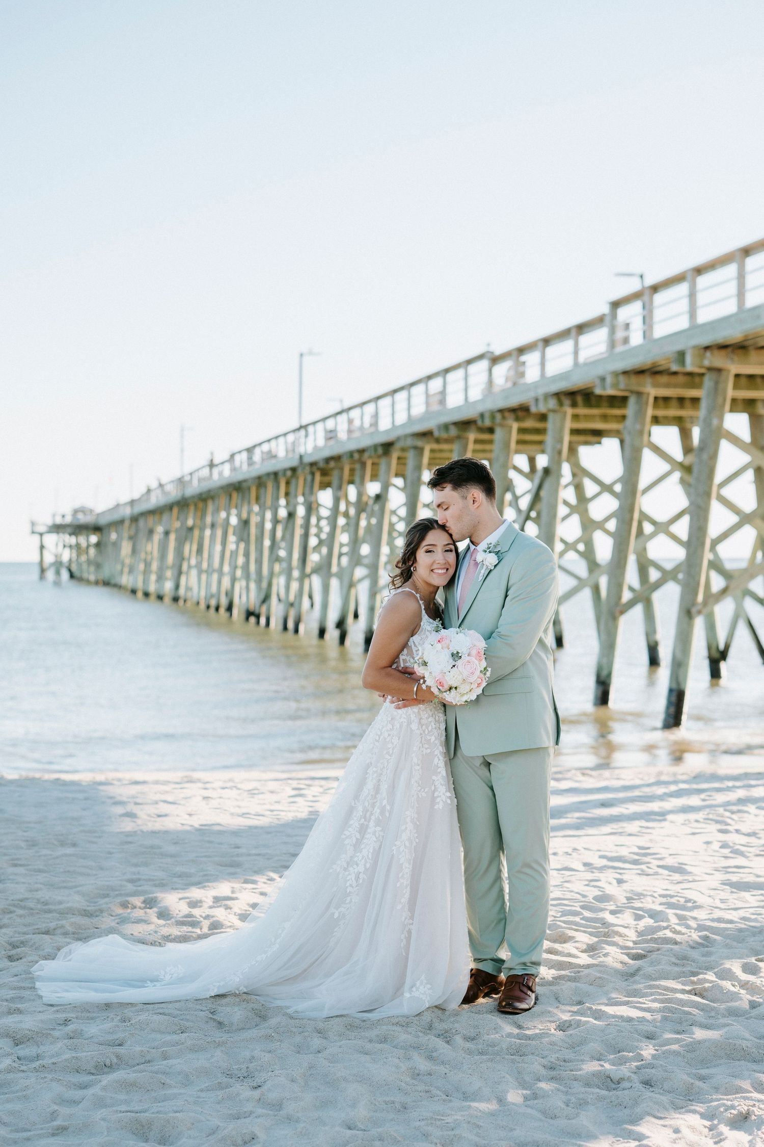 A couple poses for wedding photos on a sandy beach with a long wooden pier in the background.