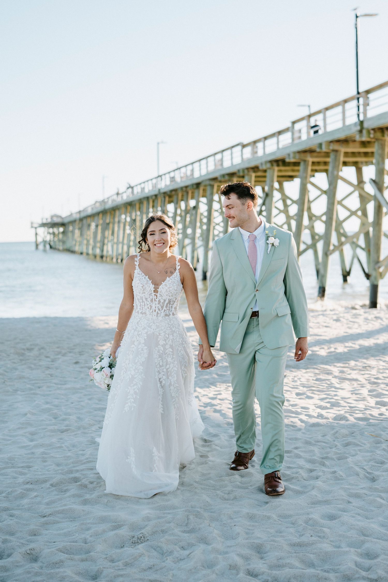 A couple walks together on a sandy beach near a wooden pier in soft pastel colors.