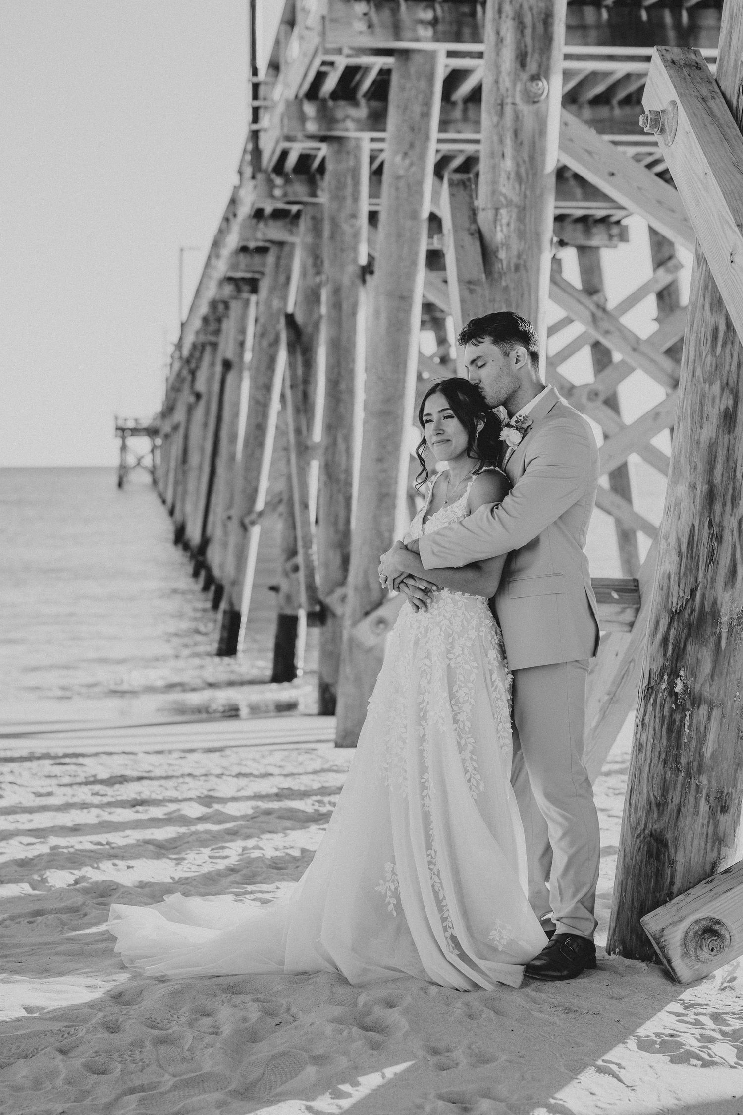 Black and white wedding portraits captured under a beach pier with dramatic lighting.
