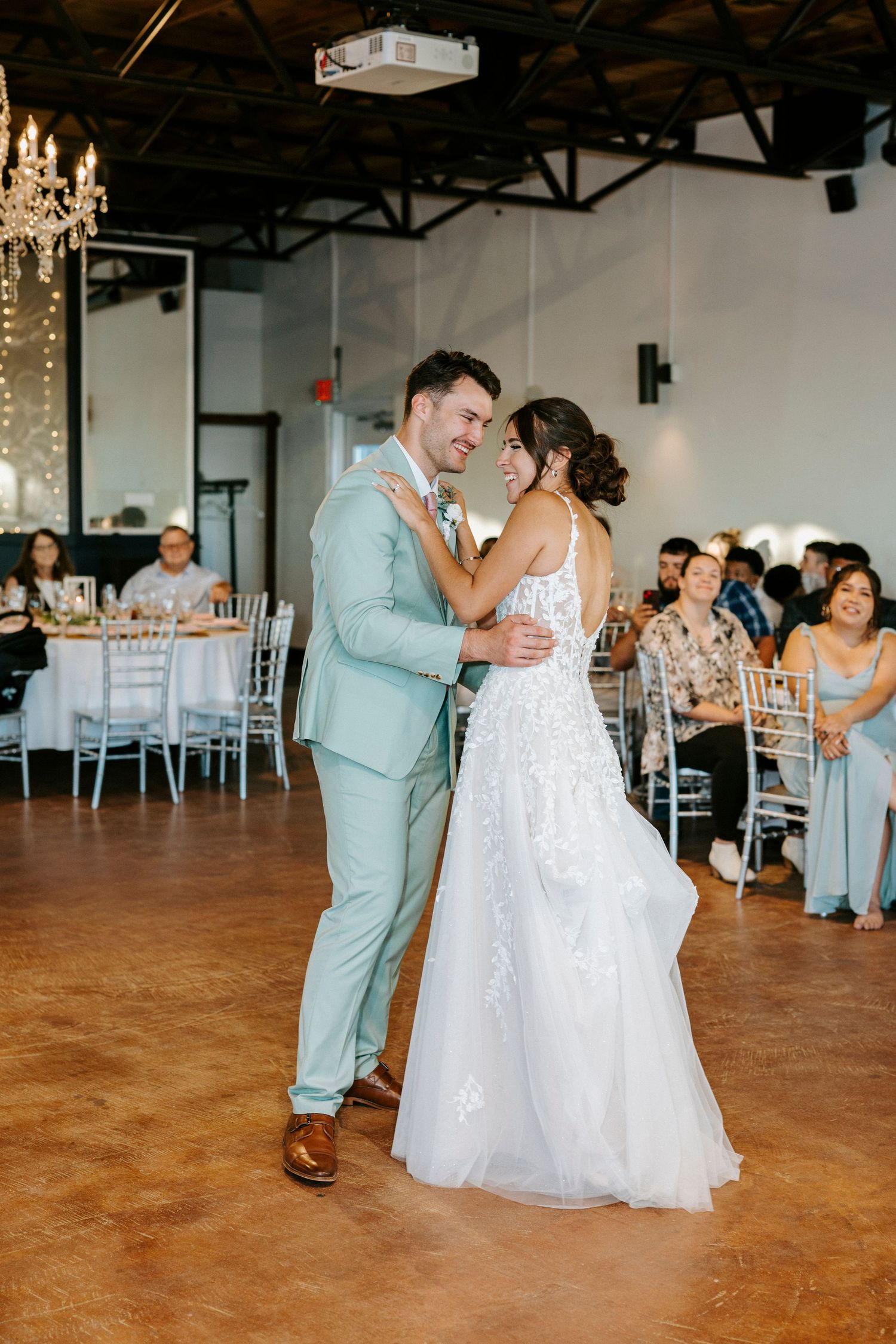 A couple shares a romantic dance on a wooden floor at their wedding reception with guests watching in the background.