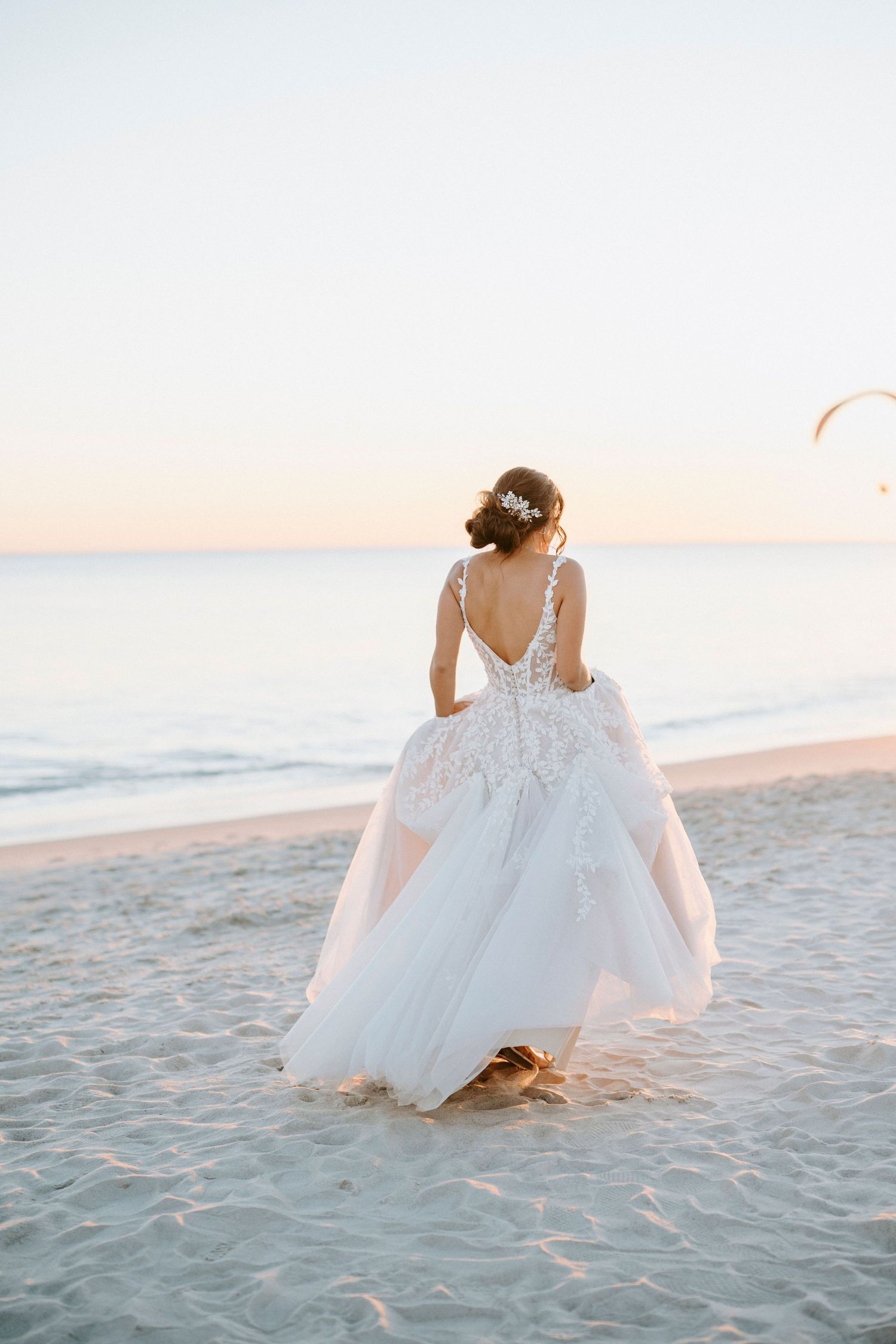A bride in a white lace wedding dress sits gracefully on a sandy beach at sunset creating a romantic coastal scene.